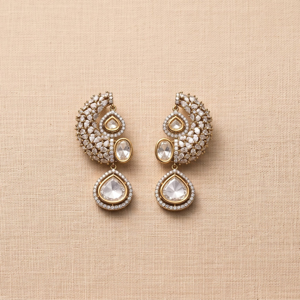 A pair of gold-toned traditional Indian earrings embellished with clear crystals and featuring teardrop-shaped clear stones, displayed on a textured beige fabric background.