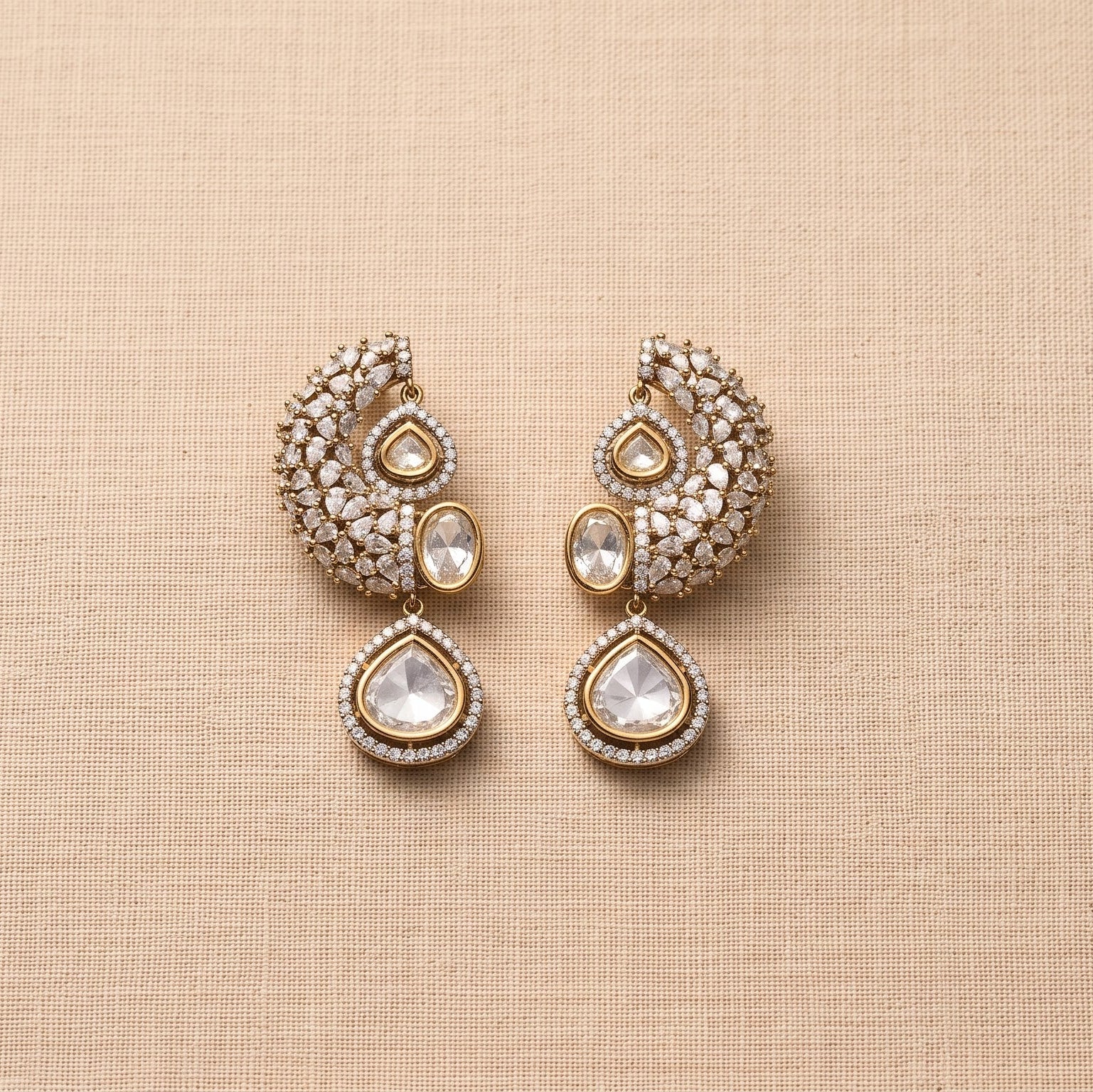 A pair of gold-toned traditional Indian earrings embellished with clear crystals and featuring teardrop-shaped clear stones, displayed on a textured beige fabric background.